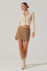 A woman is wearing a cream-colored, long-sleeve, button-up cardigan with a textured, open-knit pattern. She pairs it with a high-waisted, A-line mini skirt in a light brown/tan color. Her footwear consists of ankle-high, beige boots. She has her hair styled in a sleek, low bun and is accessorized with gold earrings.